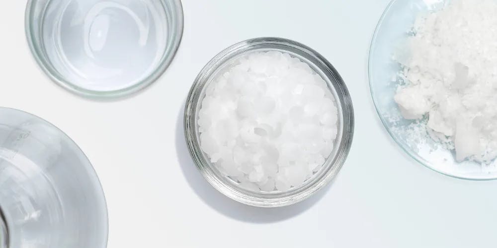 Three transparent laboratory dishes, one containing white pellets, and the other two contain a powdery cosmetic ingredients.