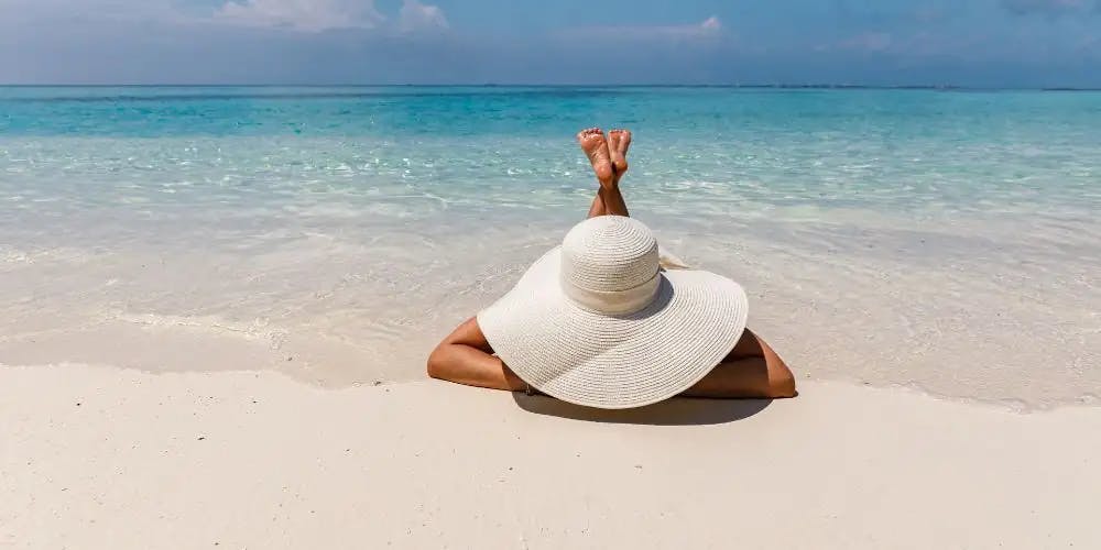 A woman in a big hat lies on her stomach on the beach and sunbathes. In the background you can see the turquoise sea.
