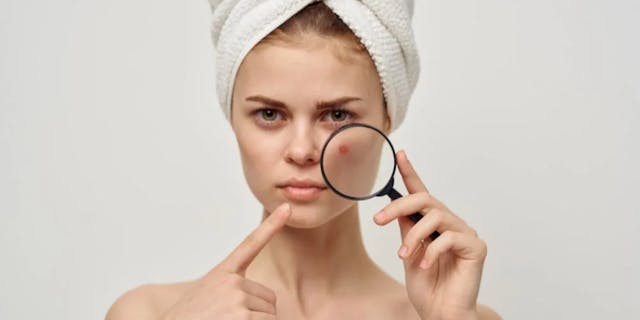 A woman with a white towel on her head examines her skin through a magnifying glass
