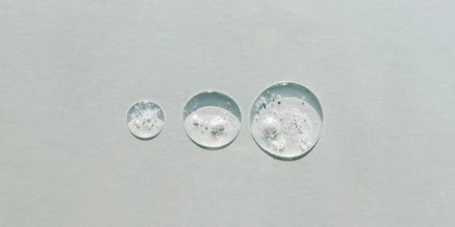 Three different sized drops of transparent serum on a gray background.