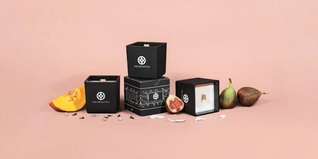 Czarne pudełka ze świecami sojowymi Nudmuses-Holibeautica, otoczone naturalnymi dodatkami jak dynia i figi na pastelowym tle.